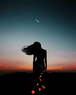 woman gazing up at crescent moon in early evening with trail of string lights behind her