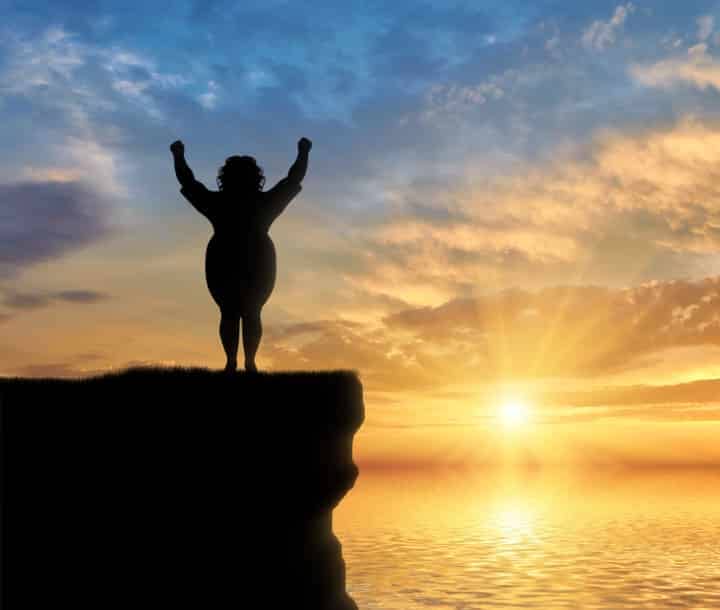overweight woman raising arms in salute to sunset