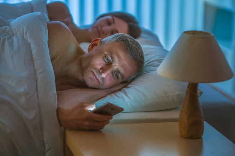sleepless man looking at phone with partner asleep next to him